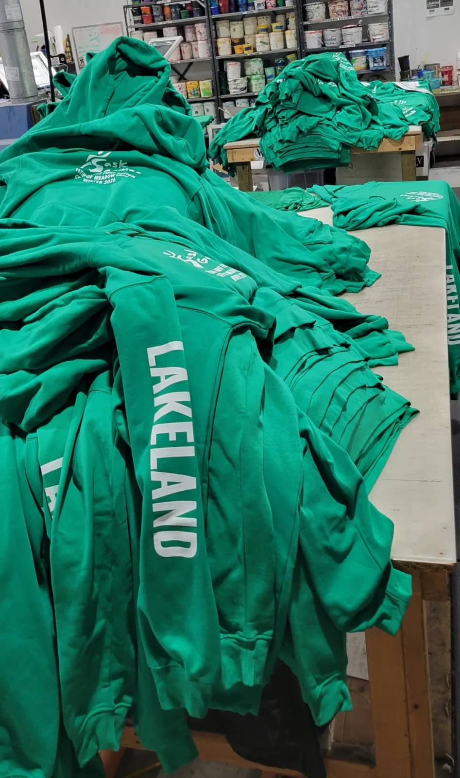 Stacks of green Sask Games hoodies with LAKELAND printed down the sleeve, piled on production tables at Sandstar Graphics