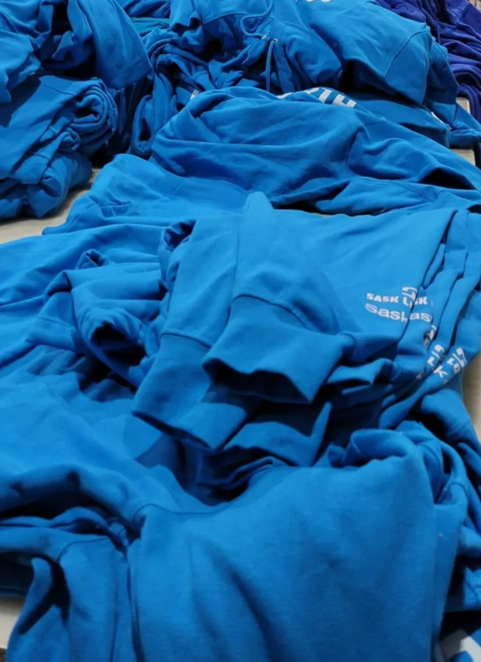 Pile of blue Sask Games team hoodies with printed sleeve text, part of a 6,000 piece order