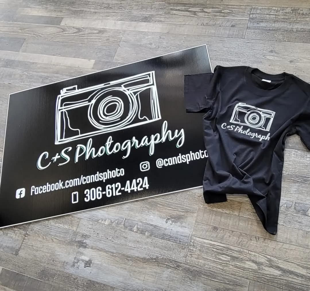 Black C+S Photography sign with camera logo and social handles beside a matching black t-shirt printed with the same logo