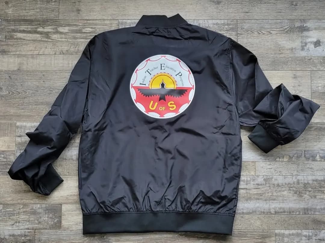 Back of a black bomber jacket with the Indian Teacher Education Program U of S logo printed in full colour