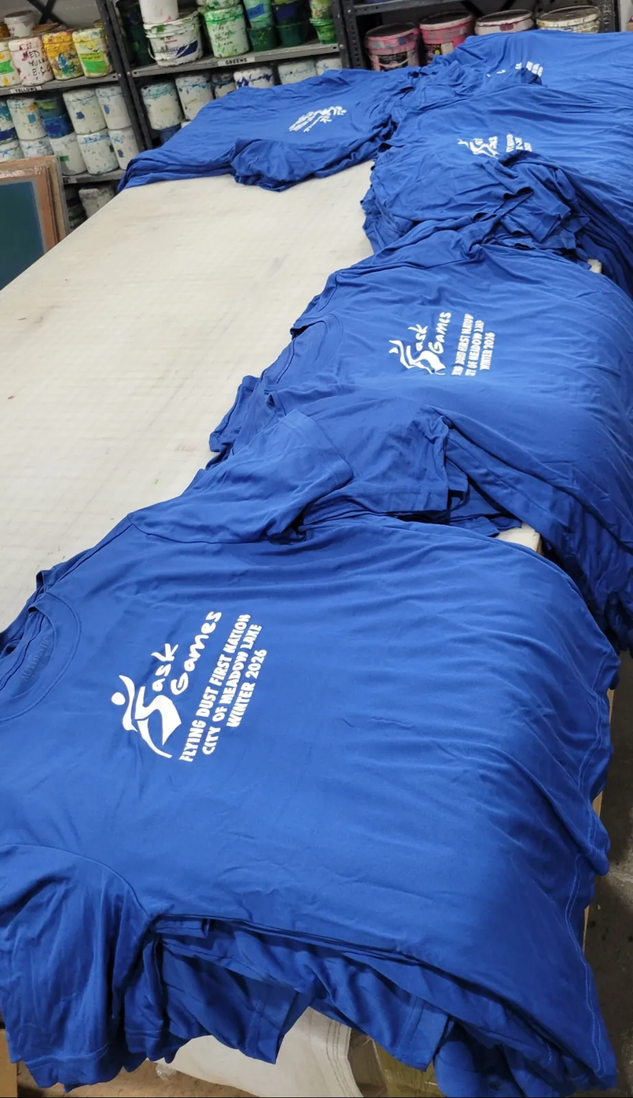 Rows of royal blue Sask Games team t-shirts printed for Winter 2026 (Flying Dust First Nation and City of Meadow Lake), laid out on a print shop table in front of shelves of ink